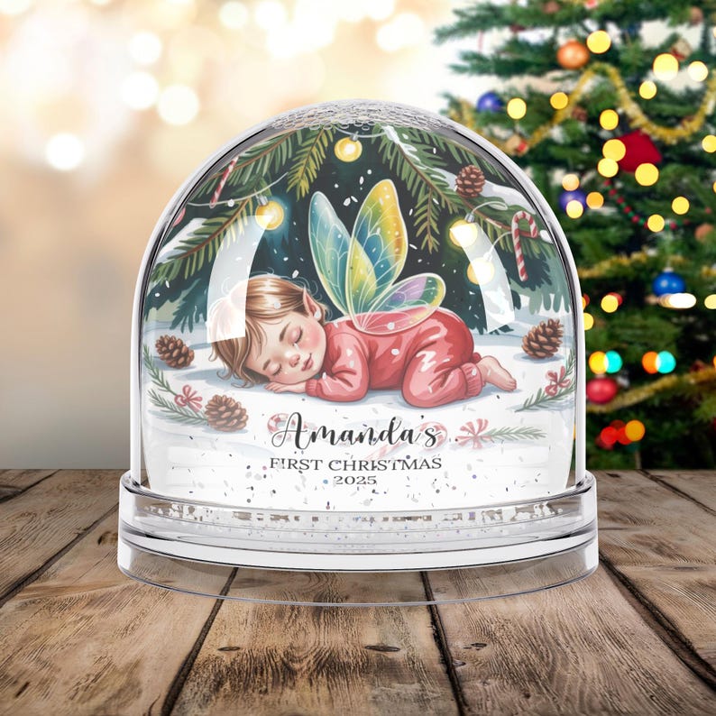 Może przedstawiać: Przezroczysta kula śnieżna ze śpiącą wr&oacute;żką w środku. Dziecko ma na sobie czerwony str&oacute;j i kolorowe skrzydła. Kula jest ozdobiona igłami sosnowymi, śniegiem i napisem "Amanda's First Christmas 2025".
