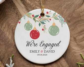 Puede incluir: Adorno navideño redondo de cerámica blanca con una cuerda dorada para colgar. Presenta un diseño festivo con bolas de Navidad verdes y rojas, bayas y el texto "We're Engaged" con nombres y una fecha.