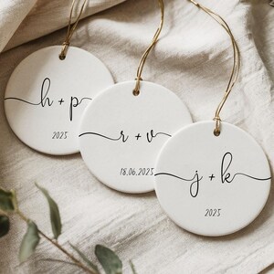 May include: Three white ceramic ornaments with gold string hangers. Each ornament features a handwritten style initial and date. The ornaments are round and have a minimalist design, perfect for personalized gifts.