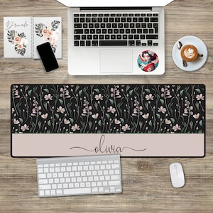 May include: A black desk mat with a floral pattern and a light pink band at the bottom. The text "olivia" is written in cursive on the band. The mat is on a wooden desk with a laptop, keyboard, mouse, and a cup of coffee.