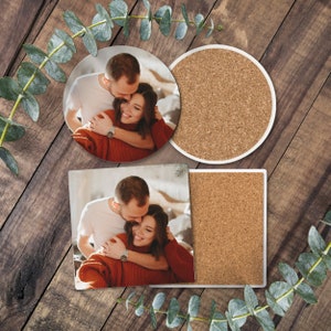 以下が含まれることがあります： 2つのコースター。1つは抱き合っているカップル、もう1つはコルクの表面。カップルは赤いセーターと白いシャツを着ています。コースターは丸みを帯びた縁のある白いものです。