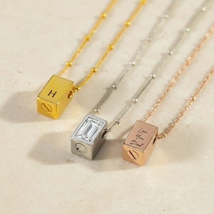 Puede incluir: Una colección de collares con colgantes cúbicos. Un collar dorado presenta la letra "H" en el colgante. Otro tiene una piedra clara rectangular. El tercer colgante es de oro rosa y tiene grabadas las palabras "Love You Dad".