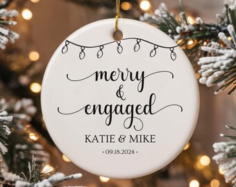 Puede incluir: Adorno navideño de cerámica blanca con texto negro que dice "merry & engaged" con los nombres Katie & Mike y la fecha 09.18.2024. El adorno está decorado con una cadena de luces.