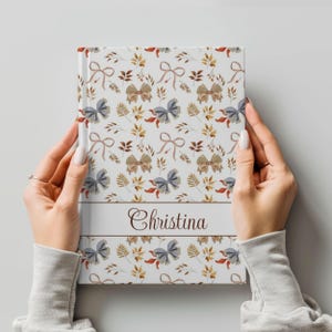 Puede incluir: Un diario blanco con un patrón floral con lazos azules y marrones, hojas y tallos. El nombre "Christina" está impreso en marrón en una pancarta azul en la parte delantera.