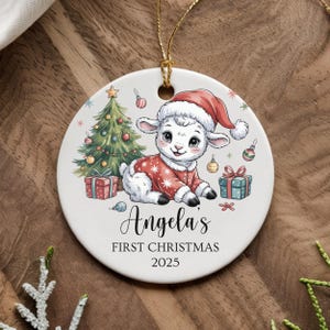 以下が含まれることがあります： サンタ帽と赤い衣装を着た子羊が描かれた丸いクリスマスオーナメント。クリスマスツリー、プレゼント、そして「Angela's FIRST CHRISTMAS 2025」の文字も表示されています。オーナメントは白で、金色の紐が付いています。