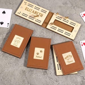 May include: A wooden cribbage board engraved with "ELIZABETH" and "EST. 202-". The board is open, showing pegging tracks and gold pegs. Three brown leather notebooks with wooden inserts are also present. The notebooks have personalized engravings.