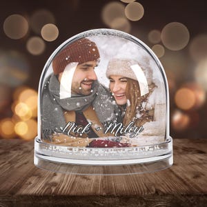 Peut inclure: Un globe à neige transparent avec une photo d'un couple souriant en vêtements d'hiver. Les noms "Nick + Miley" sont écrits en cursive sur la photo. Le globe repose sur une base transparente, posé sur une surface en bois.