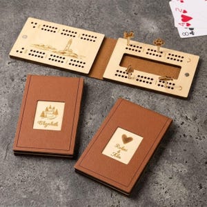 May include: Wooden cribbage board with a lighthouse design and two brown leather-bound card holders. One card holder has the name "Elizabeth" engraved, and the other has "Robert & Alex" with a heart. Includes wooden pegs.