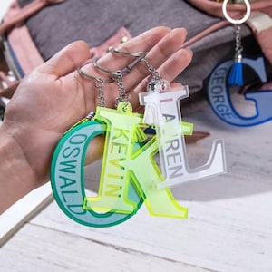 May include: Three acrylic keychains with the names "Lauren", "Kevin", and "Oswald" written on them. The keychains are in the shape of the first letter of each name. The "Lauren" keychain is clear with white lettering, the "Kevin" keychain is yellow with green lettering, and the "Oswald" keychain is green with white lettering.