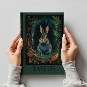 May include: A hardcover journal with a forest scene featuring a rabbit. The cover is dark green with the name "TAYLOR" in white. The illustration shows a brown and gray rabbit in a forest setting with flowers and ferns.