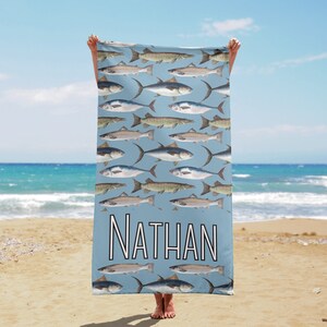 Puede incluir: Una toalla de playa azul claro con un patrón de ilustraciones de peces. La palabra "NATHAN" está impresa en letras blancas en la parte inferior. La toalla se sostiene contra un fondo de playa y océano.