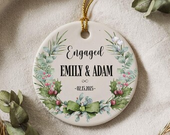 Puede incluir: Adorno redondo de cerámica blanquecina con la palabra "Engaged" en escritura negra. Los nombres "EMILY & ADAM" están centrados debajo, con la fecha "02.15.2025" debajo. Decorado con follaje verde, bayas rojas y un lazo verde.