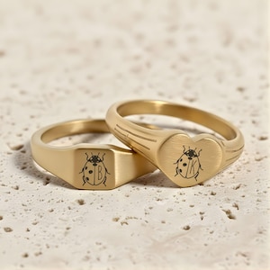 Könnte beinhalten: Zwei goldfarbene Siegelringe. Ein Ring hat eine quadratische Oberfläche mit einem Marienkäfer-Design und dem Buchstaben "B". Der andere Ring hat eine herzförmige Oberfläche mit einem Marienkäfer-Design und dem Buchstaben "K".