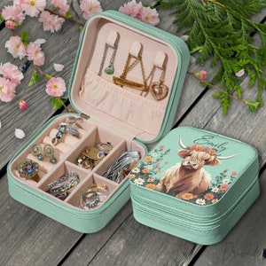 Peut inclure: Une boîte à bijoux de voyage vert menthe, ouverte pour afficher bagues, boucles d'oreilles et colliers. Le couvercle présente une illustration de vache des Highlands avec le nom "Emily" et des motifs floraux. La boîte est zippée et a un intérieur rose clair.