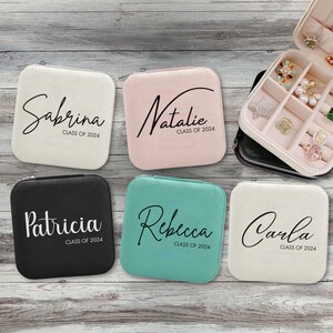 May include: Five small square jewelry boxes with different colored fabric covers. Each box has a name and "Class of 2024" printed on the front. The names are Sabrina, Natalie, Patricia, Rebecca, and Carla.