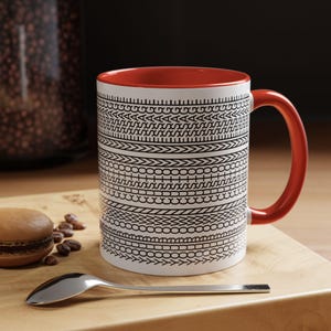 Könnte beinhalten: Weißer Kaffeebecher mit rotem Innenraum und Henkel. Der Becher hat ein schwarzes geometrisches Muster. Ein silberner Löffel und ein Macaron liegen auf einer Holzoberfläche neben der Tasse.