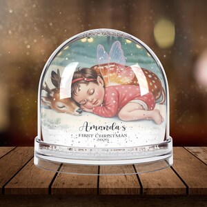 K&ouml;nnte beinhalten: Eine Schneekugel mit einem schlafenden Baby mit Feenfl&uuml;geln, das auf einem Rehkitz ruht. Das Baby tr&auml;gt einen rosa Anzug und ein rotes Stirnband. Der Text "Amanda's First Christmas 2025" ist auf der Schneekugel zu sehen.