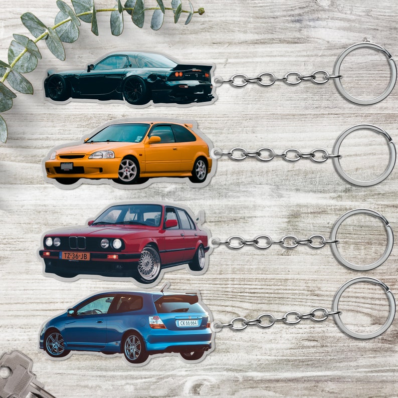 May include: Four different car keychains with silver chains and rings. The keychains feature different car models, including a black sports car, a yellow sedan, a red coupe, and a blue hatchback. The cars have license plates with the text "TZ 36-JB" and "CX 66 844".