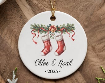 Puede incluir: Un adorno navideño redondo con una cuerda dorada. El adorno blanco presenta dos calcetines navideños rojos y blancos, una guirnalda verde y cintas rojas. Los nombres "Chloe & Noah" y el año "2025" están impresos en el adorno.