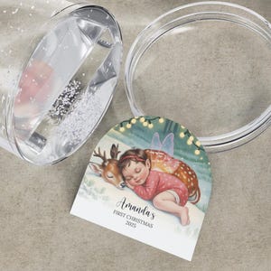 K&ouml;nnte beinhalten: Ein klarer Becher im Schneekugel-Stil mit Deckel und einem dekorativen Anh&auml;nger. Der Anh&auml;nger zeigt eine Illustration eines schlafenden Baby-Feen und Rehs mit dem Text "Amanda's FIRST CHRISTMAS 2025". Der Becher ist teilweise mit Glitzer gef&uuml;llt.
