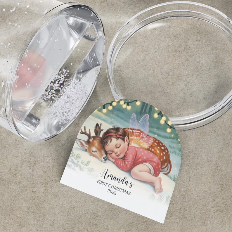 K&ouml;nnte beinhalten: Ein klarer Becher im Schneekugel-Stil mit Deckel und einem dekorativen Anh&auml;nger. Der Anh&auml;nger zeigt eine Illustration eines schlafenden Baby-Feen und Rehs mit dem Text "Amanda's FIRST CHRISTMAS 2025". Der Becher ist teilweise mit Glitzer gef&uuml;llt.