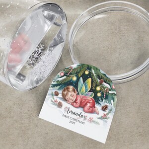 Może przedstawiać: Przezroczysta kula śnieżna z ozdobną etykietą. Etykieta przedstawia śpiącą wr&oacute;żkę w czerwonym stroju z napisem "Amanda's First Christmas 2023". Kula jest częściowo wypełniona białym brokatem i r&oacute;żowym przedmiotem.