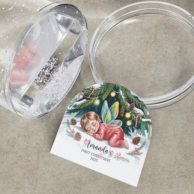 Może przedstawiać: Przezroczysta kula śnieżna z ozdobną etykietą. Etykieta przedstawia śpiącą wr&oacute;żkę w czerwonym stroju z napisem "Amanda's First Christmas 2023". Kula jest częściowo wypełniona białym brokatem i r&oacute;żowym przedmiotem.