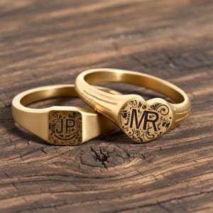 Op de afbeelding: Twee goudkleurige zegelringen. Eén ring heeft een vierkant vlak met de initialen "JP" en decoratief krulwerk. De andere ring heeft een hartvormig vlak met de initialen "MR" en vergelijkbaar krulwerk. Beide ringen liggen op een houten oppervlak.