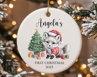 Può includere: Ornamento natalizio rotondo in ceramica bianca con cordino dorato. Presenta una volpe festosa in costume da Babbo Natale, un albero di Natale decorato e la scritta "Angela's First Christmas 2025".