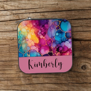 May include: A small square zippered pouch with a colorful abstract design featuring swirls of pink, blue, yellow, and purple. The pouch has a pink bottom with the name "Kimberly" printed in black.