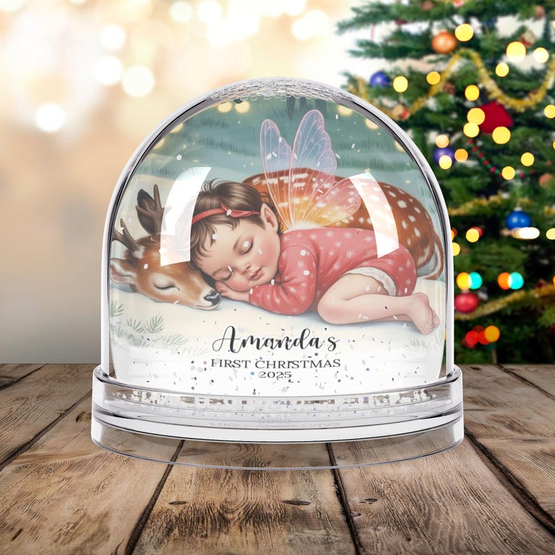 K&ouml;nnte beinhalten: Eine klare Schneekugel mit einem schlafenden Baby-Fee und einem Reh im Inneren. Das Baby tr&auml;gt einen rosa Anzug und hat Feenfl&uuml;gel. Der Text "Amanda's First Christmas 2025" ist auf der Basis aufgedruckt.
