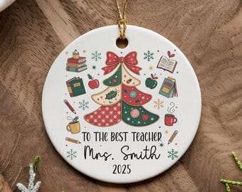 May include: A white ceramic Christmas ornament with a gold string. The ornament features a Christmas tree design with books, apples, and a bow. Text reads "TO THE BEST TEACHER Mrs. Smith 2025".