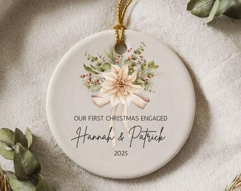 Puede incluir: Un adorno navideño de cerámica blanca con una ilustración de acuarela de una flor de pascua y vegetación. El adorno dice "Our First Christmas Engaged Hannah & Patrick 2025".
