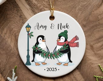 May include: A round, white ceramic Christmas ornament with a gold string. The ornament features two cartoon penguins holding hands, with a street lamp on the left. Text reads "Amy & Nick" and "2025". One penguin wears a green hat and red scarf.