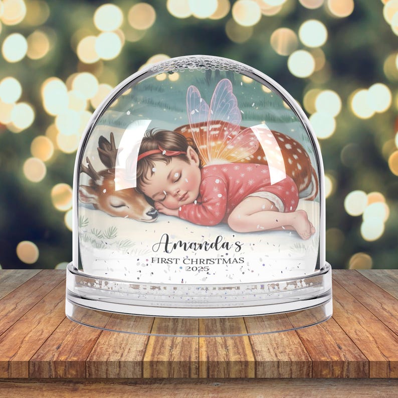 K&ouml;nnte beinhalten: Eine klare Schneekugel mit einem schlafenden Baby-Fee und einem Rehkitz darin. Das Baby tr&auml;gt einen rosa Anzug und hat Feenfl&uuml;gel. Der Text "Amanda's First Christmas 2025" ist auf der Basis aufgedruckt. Die Schneekugel steht auf einer Holzoberfl&auml;che.