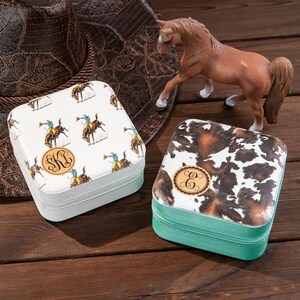 May include: Two square jewelry boxes with different designs. One box has a white background with a pattern of cowboys riding horses and a monogram "SKL". The other box has a white background with a brown and white cowhide pattern and a monogram "E".