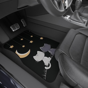 May include: Black car floor mats with a whimsical design featuring two cartoon cats gazing at a crescent moon and stars. The mats are placed in a car interior, near the pedals and seat.