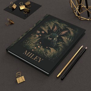 May include: A black journal with a gold-colored name "MILEY" on the front cover. The cover features a detailed illustration of a rabbit in a forest setting with ferns and flowers.