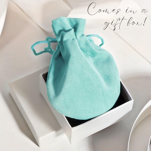 May include: A light blue velvet pouch is inside a white gift box. The box is open and the pouch is visible. The text "Comes in a gift box!" is written in cursive above the box.