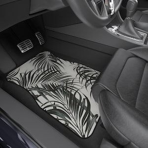 May include: Car floor mat with a white background and a pattern of dark green palm leaves. The mat is rectangular with a black border and is placed in the driver's side footwell of a car. The car interior is black.