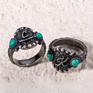 May include: Two silver rings with turquoise stones and engraved letters. The ring on the left has the letter "C" engraved on it, and the ring on the right has the letter "K" engraved on it.
