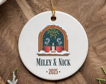 May include: A round ceramic Christmas ornament with a gold string. The ornament features a window scene with two red mugs, a lit candle, and the text "MILEY & NICK • 2025."