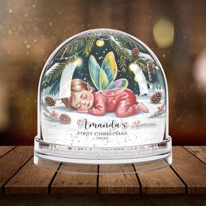Może przedstawiać: Przezroczysta kula śnieżna ze śpiącą wr&oacute;żką w środku. Dziecko ma na sobie czerwony str&oacute;j i kolorowe skrzydła. Kula jest ozdobiona igłami sosnowymi, światełkami i napisem "Amanda's First Christmas 2025".