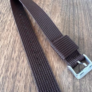 Könnte beinhalten: Ein dunkelbraunes Uhrenarmband mit gewebter Textur. Das Armband hat eine transparente, rechteckige Schnalle. Das Armband liegt auf einer Holzoberfläche.