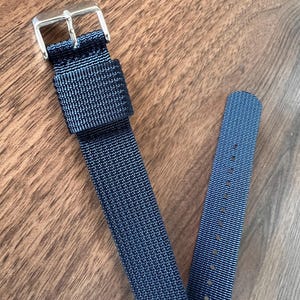 Könnte beinhalten: Ein marineblaues Uhrenarmband mit einer silbernen Schnalle. Das Armband besteht aus gewebtem Material und hat mehrere Löcher zur Anpassung. Der Hintergrund ist eine Holzoberfläche.