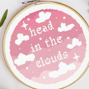 Op de afbeelding: Een roze en witte borduurring met de tekst "head in the clouds" omringd door witte wolken.
