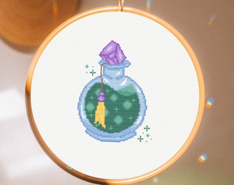 Peut inclure: Un cerceau de broderie en point de croix avec une fiole de potion violette et verte avec un pompon jaune. La fiole est entourée d'étoiles blanches.