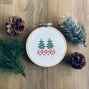 Peut inclure: Un cerceau en bois pour broderie avec un tissu blanc à l'intérieur. Le tissu présente un motif de point de croix de deux arbres verts avec des accents rouges. Le cerceau est entouré de branches de pin et de pommes de pin.
