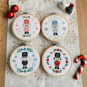 May include: Four round embroidery hoops with different designs of nutcracker soldiers. Each hoop has a white background with colorful embroidery of a nutcracker soldier in a different pose and color scheme. The hoops are arranged in a square shape on a white fabric background with a snowflake pattern.