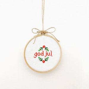 May include: A white embroidery hoop with a wooden frame, hanging from a twine string. The embroidery reads "god jul" in red lettering with a green wreath around it.